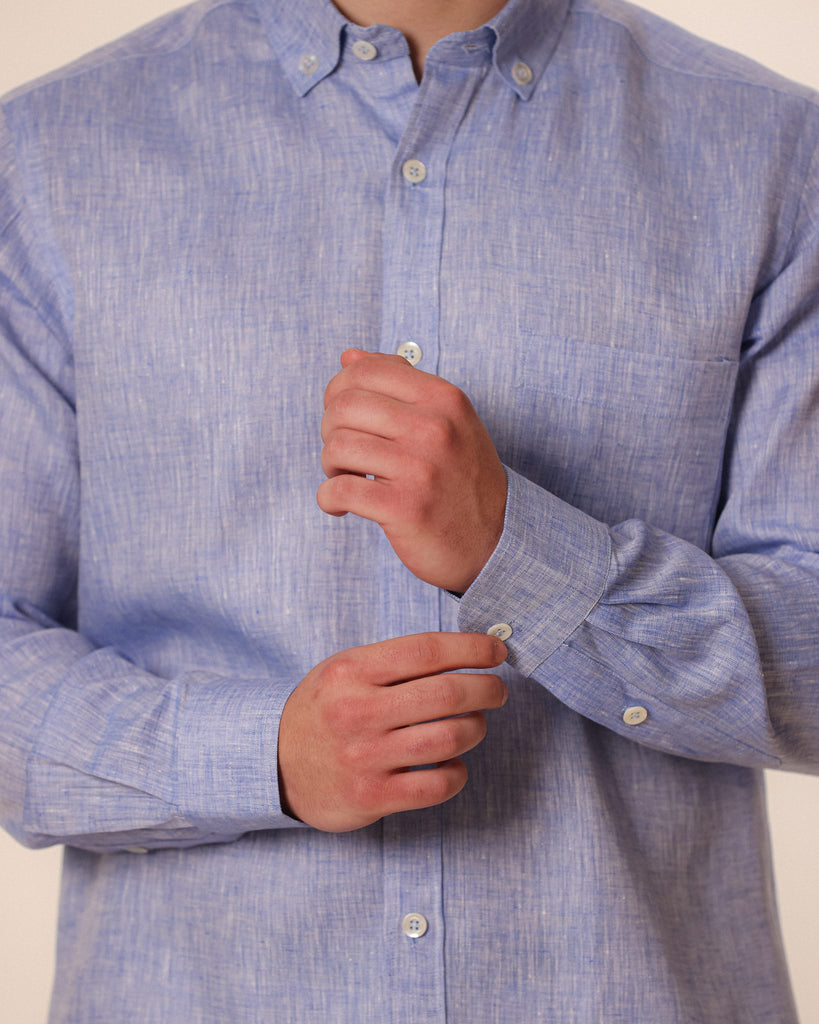 A close-up of a man wearing a light blue textured linen button-down shirt, adjusting the cuff with one hand while buttoning the other cuff.